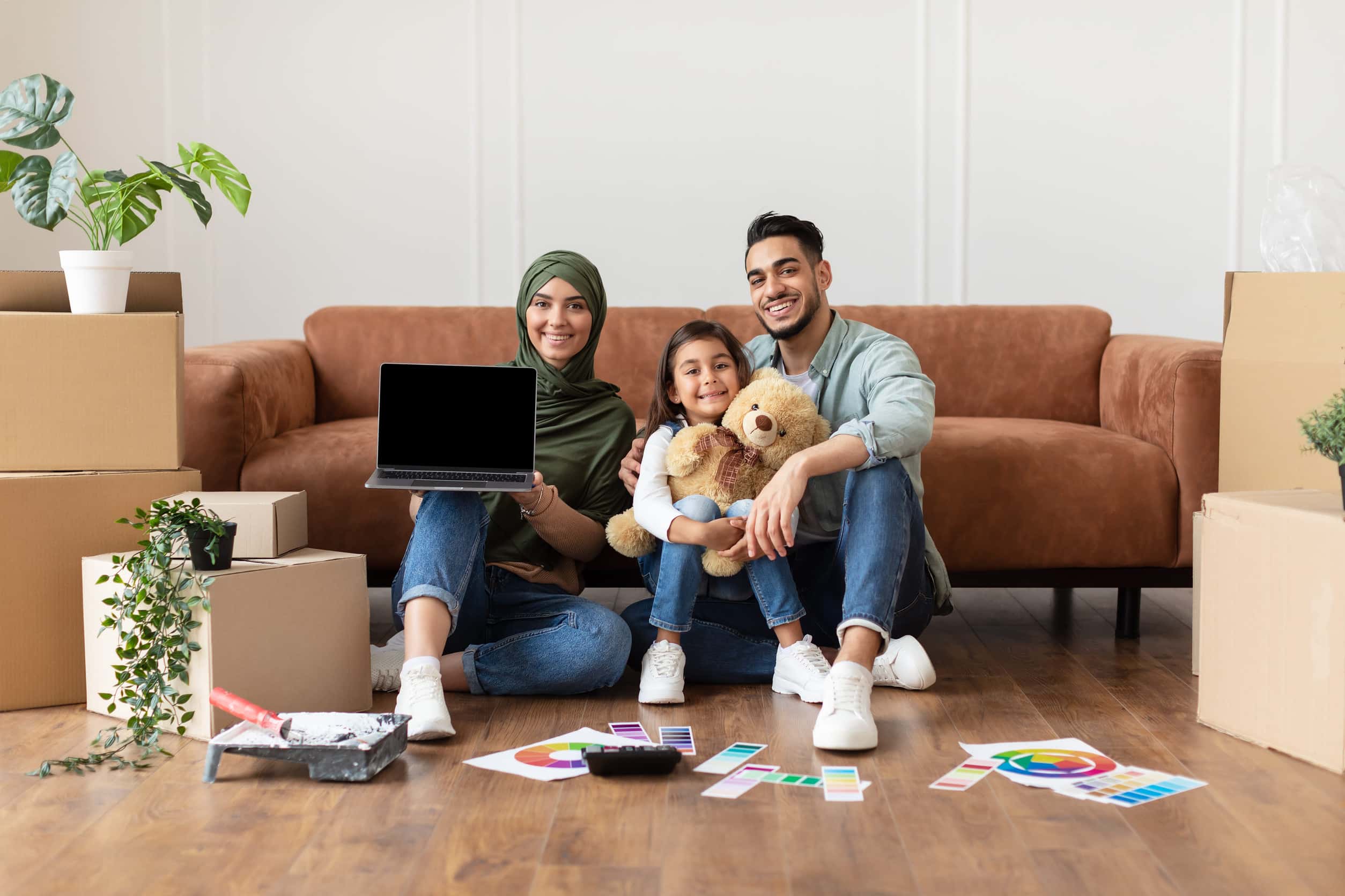 A young family sitting among moving boxes in their new home, using a laptop, paint swatches, and a calculator to plan and budget their move